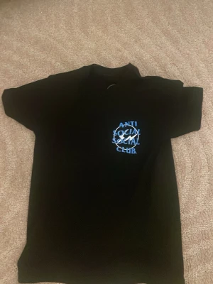 Svart Anti Social Social Club t-shirt - Säljer en svart t-shirt från Anti Social Social Club med blå och vit logga på bröstet. T-shirten är i 100% bomull och har en klassisk passform med korta ärmar. Perfekt för dig som gillar streetwear och vill ha något stilrent men ändå med edge.