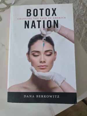 Botox Nation av Dana Berkowitz - En bok med vit framsida och ett foto av en kvinna som får en spruta i pannan. Titeln är 'Botox Nation: Changing the Face of America' och författaren är Dana Berkowitz. Boken undersöker botoxkulturens påverkan på samhället och skönhetsideal.