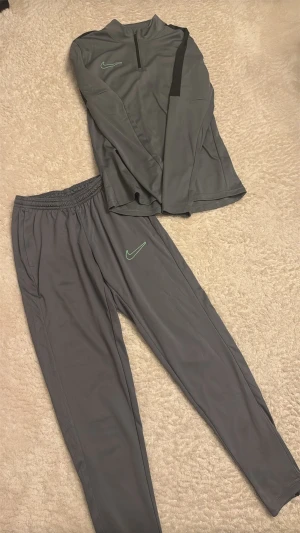 Grått Nike träningsset - Säljer ett grått träningsset från Nike med långärmad tröja och matchande byxor. Tröjan har halv dragkedja och svart detalj på axeln, medan byxorna har resår i midjan. Båda plaggen har Nike-logga i ljusgrönt. Perfekt för träning eller chill.