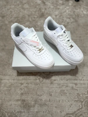 Vita Nike Air Force 1 sneakers - Klassiska helvita Nike Air Force 1 sneakers i lågt utförande. Skorna har en stilren design med perforerad tå, vita skosnören och den ikoniska swoosh-loggan på sidan. Ovandelen är i slätt läder och sulan är tjock och vit för extra komfort.
