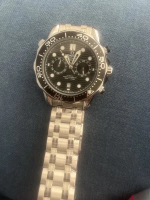 Omega Seamaster klocka i silver - Stilren Omega Seamaster klocka med svart urtavla och silverfärgad länk i metall. Klockan har tydliga markeringar, kronograffunktion och vridbar bezel. Extra länkar medkommer!