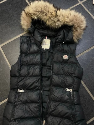 Svart dunväst med pälskant Moncler - Snygg svart dunväst från Moncler med avtagbar huva och fluffig pälskant. Västen har två dragkedjefickor fram och klassisk Moncler-logga på bröstet. Perfekt för lager på lager och håller dig varm med stil.