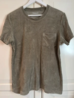 Grön frotté t-shirt från Kappahl - Säljer en grön t-shirt från Kappahl i mjuk frotté. T-shirten har korta ärmar, rund halsringning och en bröstficka som detalj. Perfekt för en avslappnad och trendig look. I storlek S.