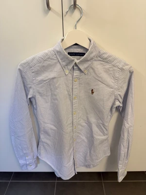 Randig skjorta från Ralph Lauren, stl XS - Klassisk vit och ljusblå randig skjorta från Ralph Lauren med broderad logga på bröstet. Skjortan har lång ärm, knappar framtill och button-down krage. Perfekt till en clean och stilren look.