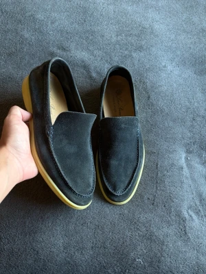 Svarta mockaloafers med gummisula - Säljer ett par stilrena svarta loafers i mocka med ljusgul gummisula. Skorna har en klassisk slip-on design utan snörning och en mjuk, rundad tå. Perfekta för dig som vill ha en clean och enkel look till vardagsoutfiten.