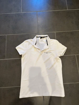 Vit piké från Sail Racing - Klassisk vit pikétröja från Sail Racing med svart logga på bröstet och svart rand på kragen. Tröjan har korta ärmar och en stilren look som passar perfekt till jeans eller shorts.