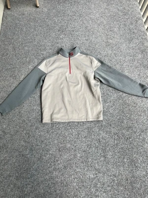 footjoy Grå halvzip tröja med ribbade ärmar - Snygg grå tröja med halvzip och röd dragkedja. Kroppen är ljusgrå medan ärmarna är mörkare grå och ribbade för extra stil. Hög krage med röd logga framtill. Perfekt till lager-på-lager och chill vardagsstil.