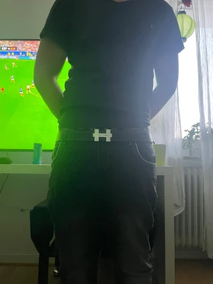 Svart bälte med silverfärgat H-spänne - Snyggt svart bälte med ett stort H-format spänne i silverfärg. Bältet har vita sömmar längs kanterna och är gjort i ett slätt material som ger en clean look. Perfekt accessoar för att lyfta vilken outfit som helst.