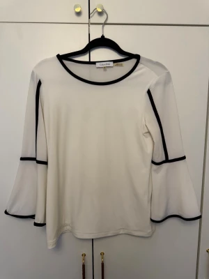 Vit långärmad topp Calvin Klein XS - Vit långärmad topp från Calvin Klein i storlek XS. Toppen har svarta kontrastkanter längs halsringning, ärmar och ärmslut. Ärmarna är utsvängda. Perfekt att styla med jeans, kjol eller kostymbyxor. 