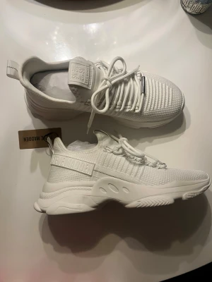 Vita Steve Madden sneakers - Snygga helvita sneakers från Steve Madden med ovandel i textil och syntetskinn, med grov sula. Skorna har rund tå, vita snören och diskret logga på sidan samt plös. Perfekta för dig som gillar en clean och modern look. (Upplevs små i storlek, innersulans mått är 26,2 cm. Helt nya aldrig använt )