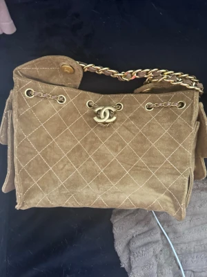 Brun mocka handväska med kedja - Snygg brun handväska i mjuk mocka med klassiskt rutmönster. Väskan har en axelrem i guld- och läderkedja samt spänne framtill. Perfekt för dig som vill ha en tidlös och trendig accessoar.