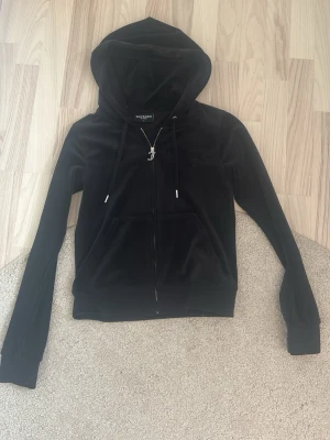 Svart juicy tröja - Säljer en svart juicy hoodie i mjuk velour med dragkedja framtill och fickor på sidorna. Tröjan har huva med snören och ribbade muddar vid ärmslut och nederkant. Perfekt för en chill och avslappnad stil.
