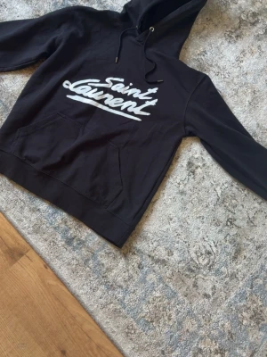 Svart Saint Laurent hoodie - Svart hoodie från Saint Laurent med stor vit logga framtill. Klassisk känguruficka och dragsko i huvan. Mjuk bomullskänsla och avslappnad passform, perfekt för en cool streetstyle-look.
