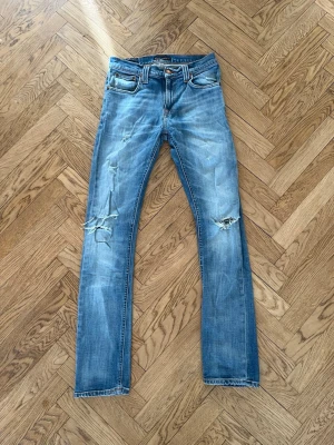 Blå slitna jeans från Nudie Jeans - Säljer ett par blå jeans från Nudie Jeans med slitningar och hål på båda knäna. Klassisk femficksmodell med raka ben och ljus tvätt. Snygga detaljer på bakfickorna och patch i läder bak i midjan. Perfekta för en avslappnad och trendig look.