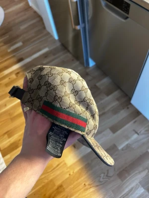 Gucci beige keps med GG-mönster - Säljer en beige keps från Gucci med det klassiska GG-monogrammet över hela kepsen. På sidan finns den ikoniska gröna och röda randen. Justerbar rem baktill och böjd skärm. Materialet är canvas med detaljer i svart. Perfekt för dig som vill ha en lyxig och trendig accessoar.