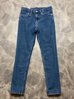 Blå slim fit jeans med hög midja - Snygga blå slim fit jeans med klassisk femficksdesign och hög midja. Jeansen har raka ben och stängs med dragkedja och knapp. Perfekta för en trendig och avslappnad stil. Materialet är klassisk denim i bomull.
