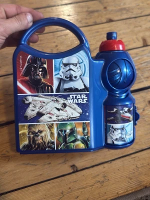 Star Wars lunchlåda med flaska ny - Blå lunchlåda i plast med Star Wars-motiv och tillhörande vattenflaska. Motiven visar Darth Vader, Stormtrooper, Millennium Falcon, Chewbacca och Boba Fett. Praktiskt handtag för enkel transport och röd kork på flaskan. Perfekt för skolan eller utflykten.