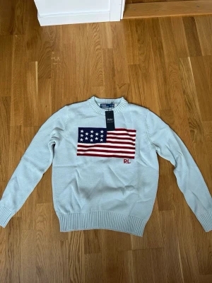 Ljusblå stickad tröja Polo Ralph Lauren - Ljusblå stickad tröja från Polo Ralph Lauren med amerikansk flagga broderad på bröstet och RL-logga i rött. Tröjan har rund halsringning, ribbade muddar och är tillverkad i bomull. Perfekt för dig som gillar klassisk preppy stil.