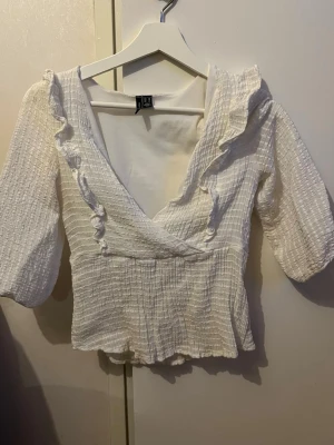 Vit omlottopp med volang från H&M - Supersöt vit omlottopp från veromoda med volangdetaljer längs ringningen och trekvartsärm. Toppen har en ribbad struktur och lätt stretchigt material som ger en bekväm passform. Perfekt att styla med jeans eller kjol för en trendig look.