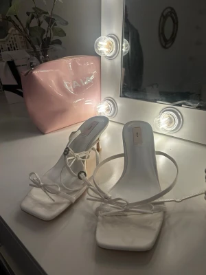 Vita sandaletter med snörning - Säljer ett par trendiga vita sandaletter med smal klack och fyrkantig tå. Skorna har tunna remmar som knyts runt foten och en elegant rosett framtill. Perfekta för dig som gillar minimalistisk och stilren design. Materialet ser ut att vara konstläder.