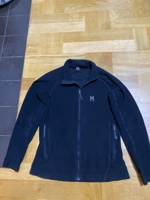 Svart fleecejacka från Haglöfs - Svart fleecejacka från Haglöfs med hel dragkedja framtill och två sidofickor med dragkedja. Jackan har hög krage och diskret logga på bröstet. Perfekt för lager-på-lager och chill dagar.