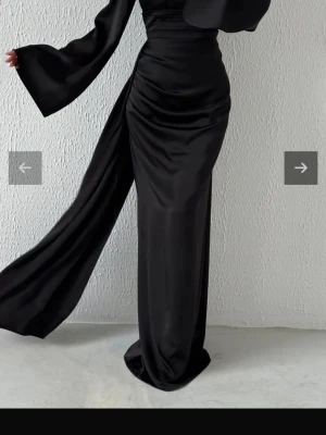 Svart långklänning med vida ärmar - Elegant svart långklänning med markerad midja och vida, långa ärmar. Klänningen har draperade detaljer och en slits framtill som ger en snygg siluett. Tillverkad i ett glansigt material som ger en lyxig känsla.