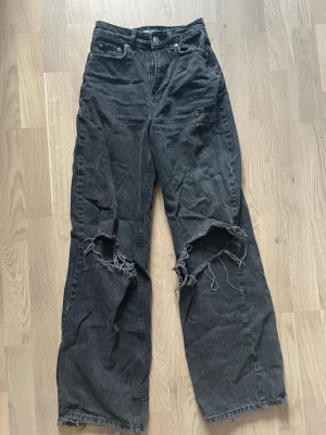 Svarta slitna jeans med vida ben - Säljer ett par svarta jeans med hög midja och vida ben. Jeansen har stora slitningar och hål vid knäna för en riktigt edgy look. Klassisk femficksmodell med dragkedja och knapp. Perfekta för dig som gillar streetstyle.