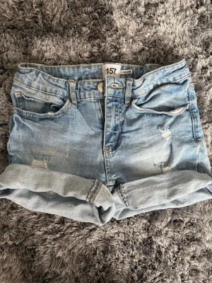 Ljusblå denimshorts från 157 - Säljer ett par ljusblå denimshorts från 157 med upprullade benslut och slitna detaljer framtill. Klassisk femficksmodell med knapp och dragkedja. Perfekta för varma dagar och enkel att matcha med allt.