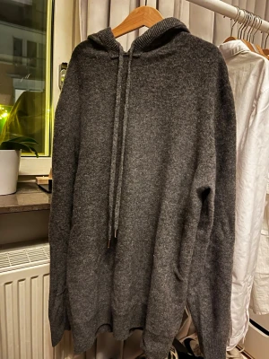 Mörkgrå hoodie i kashmire  - Mysig mörkgrå hoodie med huva och dragsko. Tillverkad i ett mjukt, stickat material som känns skönt mot huden. Hoodien har en avslappnad passform och ribbade muddar vid ärmslut och nederkant. Perfekt för chill dagar eller när du vill ha något bekvämt. Om du är sugen är det bara att höra av dig!