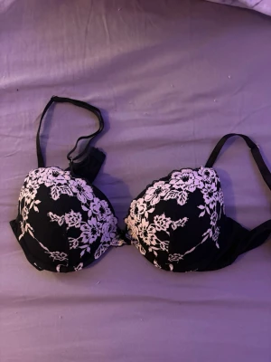 Svart push-up bh med spets från Lindex - Snygg svart push-up bh från Lindex med vita spetsdetaljer i blommigt mönster över kuporna. Justerbara axelband och bygel för extra stöd. Märkt med modellen Malva. Perfekt för dig som gillar feminina detaljer.