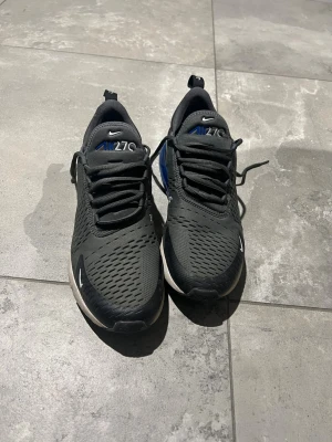 Nike Air Max 270 svarta sneakers - Nike Air Max 270 sneakers i svart mesh med blå detaljer och vit sula. Skorna har en synlig Air-enhet i hälen och klassisk Nike-logga på sidan. Snygg sportig design med snörning och ventilerande material.