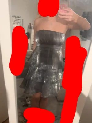 Axelbandslös silverklänning med volanger - Axelbandslös kort klänning i glittrigt silvermaterial med flera lager volanger. Klänningen har en snygg, festlig look och är perfekt för dig som vill sticka ut. Materialet är troligtvis polyester och ger ett skimrande intryck. Kan mötas upp i Stockholm 🥰