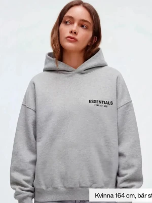 Grå hoodie Essentials Fear of God - Snygg grå hoodie från Essentials Fear of God med svart logga på bröstet och stor text på ryggen. Den är i färgen Light Heather grey och är i storlek xxs men har en lite Oversized passform, mjukt material och klassisk huva. Perfekt för en avslappnad streetwear-look.