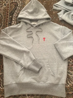 Ami paris Hoodie  - Säljer en stilren grå hoodie från Ami Paris med den ikoniska röda hjärtloggan på bröstet. Hoodien har huva med dragsko, magficka och ribbade muddar. Perfekt för dig som gillar enkel men snygg streetwear. Materialet är mjukt och skönt bomullstyg.