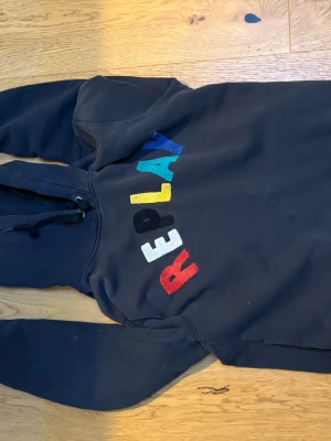 Svart hoodie från Replay med färgglatt tryck - Snygg svart hoodie från Replay med stort REPLAY-tryck i olika färger på bröstet. Hoodien har huva med dragsko och är tillverkad i mjukt material, perfekt för en avslappnad stil. Passar dig som gillar streetwear och vill sticka ut med färgglada detaljer.