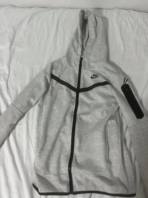 Ljusgrå Nike hoodie med dragkedja - Snygg ljusgrå hoodie från Nike med svart dragkedja och svarta detaljer. Klassisk huva, fickor på sidorna och en extra ficka med dragkedja på ärmen. Perfekt för en sportig och avslappnad stil. Tillverkad i mjukt material för extra komfort.