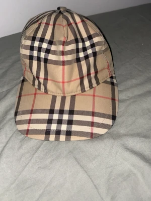 Burberry keps med klassiskt rutmönster - Snygg keps från Burberry med det ikoniska beigea rutmönstret och svart justerbar rem bak. Märkeslapp med texten 'Burberry London England' baktill. Perfekt accessoar för en trendig streetstyle-look.