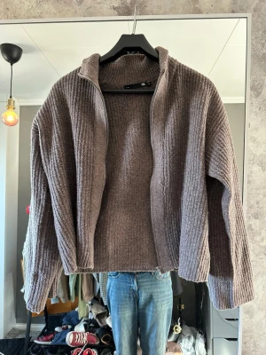 Brun ribbad ziptröja från 157 - Mysig brun ribbstickad ziptröja från 157 med hög krage och dragkedja framtill. Tröjan har långa ärmar och är tillverkad i ett mjukt, tjockt material som passar perfekt för kyligare dagar. Enkel och stilren design som är lätt att matcha.