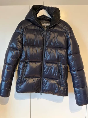 Blå pufferjacka save the duck - Snygg pufferjacka med glansig finish och huva. Jackan har två dragkedjeförsedda fickor framtill och quiltad design med breda paneler. Perfekt för kalla dagar och ger en modern look.