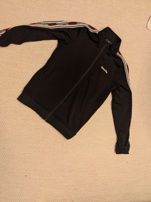 Svart track jacket från Russell - Svart track jacket från Russell med dragkedja framtill och ståkrage. Ränder i vitt och vinrött med logga längs ärmarna ger en sportig vibe. Perfekt för en avslappnad streetstyle-look.