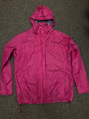 Rosa regnjacka från Didriksons - Snygg rosa regnjacka från Didriksons med huva och dragsko i nederkant. Jackan har hel dragkedja framtill med vindslå, två sidofickor med dragkedja och justerbara ärmslut. Perfekt för regniga dagar och enkel att packa ner.