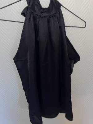 Svart halterneck topp med rosett - Snygg svart halterneck topp i glansigt material med breda axelband som knyts i en stor rosett i nacken. Toppen har lös passform och ärmlös design, perfekt till fest eller sommaren, storlek s