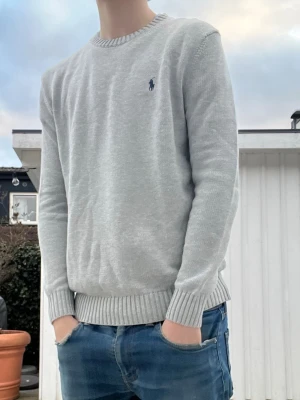 (Helt ny)Ljusgrå stickad tröja från Polo Ralph Lauren - (Moddelen är 187 och väger 77 kg )Snygg ljusgrå stickad tröja från Polo Ralph Lauren med rund halsringning och diskret blå logga broderad på bröstet. Tröjan har ribbade muddar vid ärmslut och nederkant, perfekt för lager på lager och enkel att matcha.