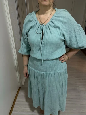 Ljusgrön klänning från Gina Tricot - Superfin ljusgrön klänning från Gina Tricot med trekvartsärm och knytning i halsen. Klänningen har en smockad midja och är gjord i ett mjukt bomullstyg som ger en avslappnad och luftig känsla. Perfekt för varmare dagar.