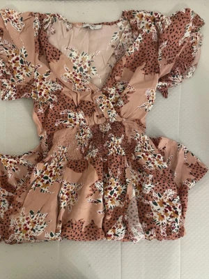 Blommig kortklänning med puffärm - Söt kortklänning i ljusrosa med blommigt och prickigt mönster. Klänningen har puffärmar, omlott över bysten och smock i midjan för en bekväm passform. Perfekt för varma dagar och sommarens alla tillfällen.