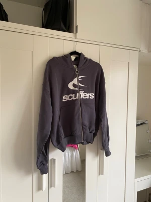 Grå hoodie med dragkedja från Scuffers - Säljer en grå hoodie från Scuffers med stor vit logga på bröstet. Hoodien har dragkedja framtill, huva och en stor magficka. Tillverkad i mjukt bomullsmaterial och har ribbade muddar vid ärmslut och nederkant. Perfekt för en avslappnad streetwear-look.