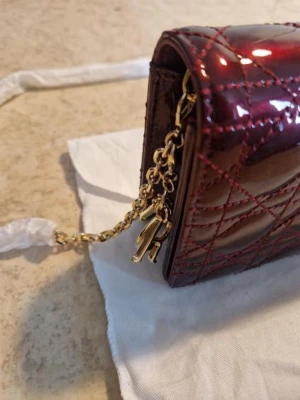 Bordeaux kuvertväska från Dior - Snygg bordeaux kuvertväska från Dior i glansigt läder med klassiskt quiltat mönster. Väskan har guldfärgad kedja och charmiga Dior-bokstäver som detalj. Magnetlås och elegant rektangulär form gör den perfekt för att lyfta din outfit.