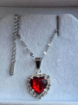 Halsband med röd hjärtformad sten - Snyggt halsband i rostfritt stål med en stor röd hjärtformad sten som hänge. Runt hjärtat sitter gnistrande klara stenar som ger extra bling. Kedjan är silverfärgad och har en justerbar längd. Perfekt för dig som vill sticka ut med ett romantiskt smycke.