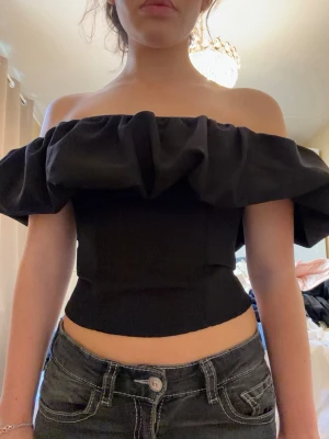 Svart offshoulder croppad topp - Säljer en svart offshoulder topp med volang runt axlarna. Toppen är croppad och har en tight passform som framhäver midjan. Perfekt för dig som vill ha en trendig och stilren look.