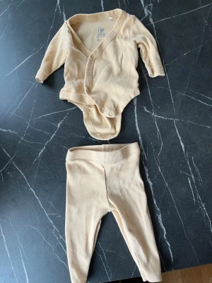 Beige ribbat babyset från KappAhl - Säljer ett mjukt och bekvämt set från KappAhl med långärmad omlottbody och matchande leggings i beige ribbad bomull. Perfekt för mysiga dagar hemma. Tryckknappar i grenen på bodyn gör det enkelt att byta blöja.